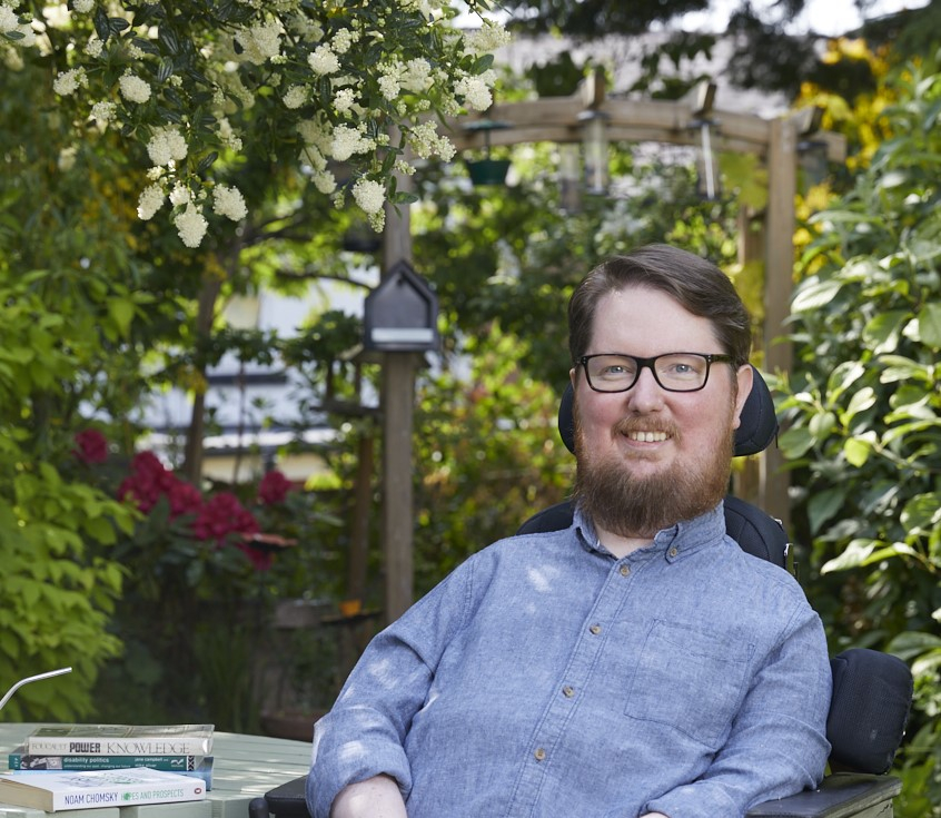 Dr Miro Griffits, a white wheelchair user with brown hair and a beard, sits in his wheelchair surrounded by a colorful garden.