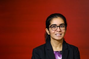 Brown-skinned woman with dark long hair wearing glasses and a black blazer looking at camera in front of a red screen.